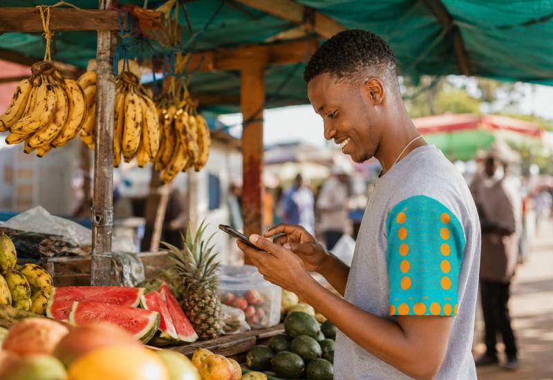 Young entrepreneur managing a fresh produce market stall while using a mobile phone to run his business in Kenya.